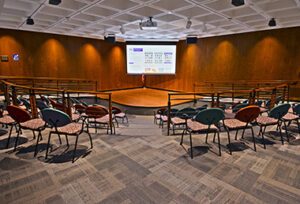 Tiered auditorium with colorful seats, wood-paneled walls, a stage, and a large projection screen.