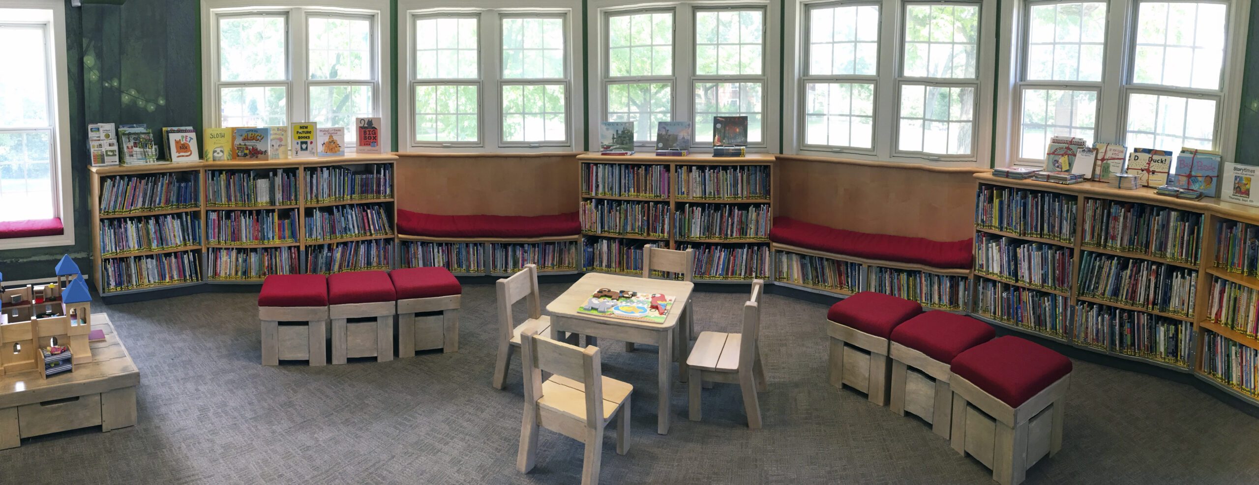 Curved bookshelves filled with books in a library reading nook designed with elements from "Journey" by Aaron Becker, featuring red cushioned seating and a small table with chairs.