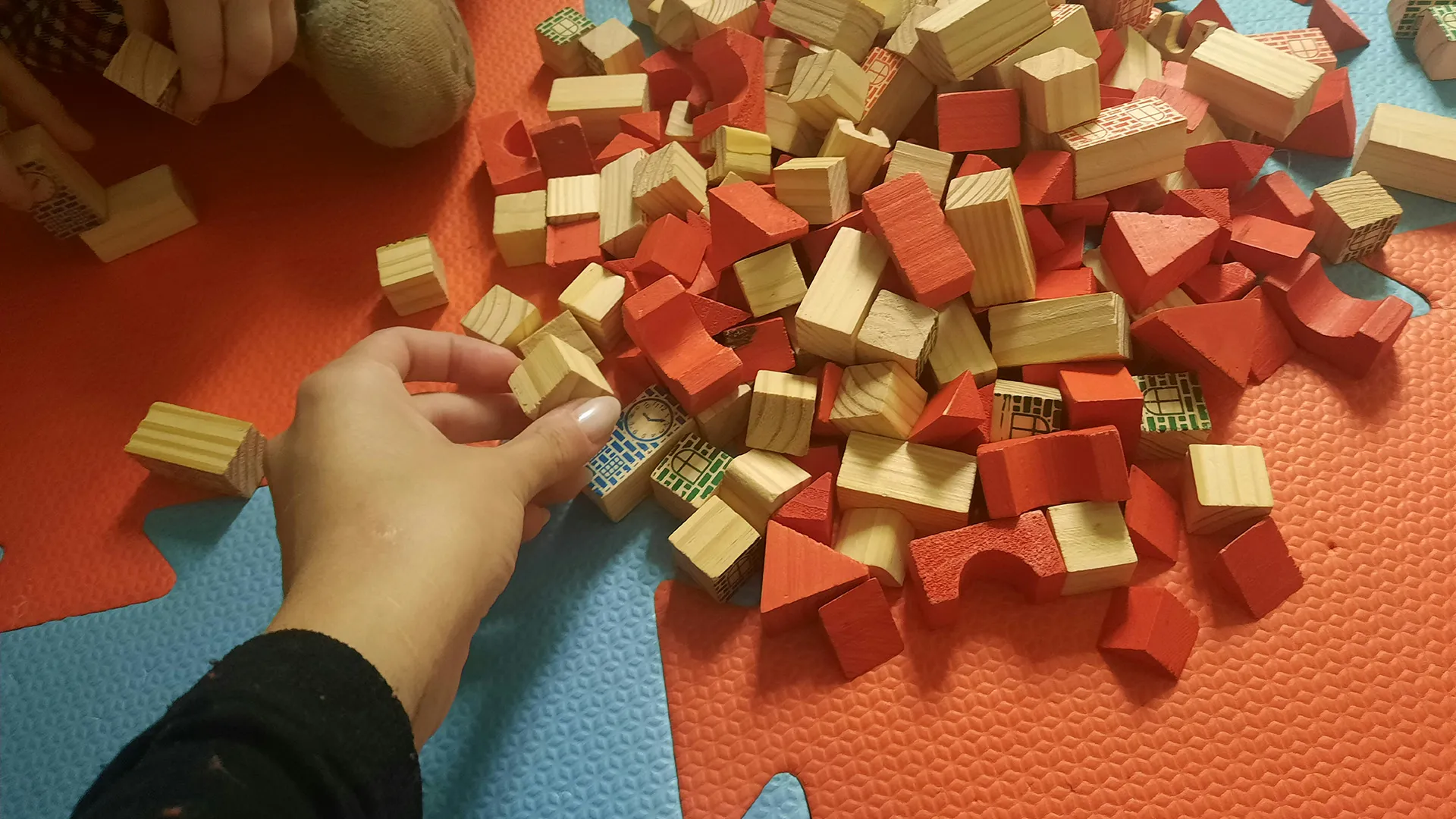 A hand takes a block from a pile of red and wooden blocks on a play mat.