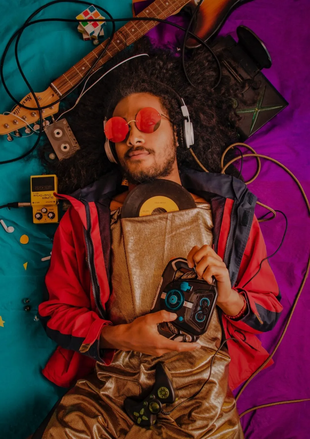 A person with curly hair, red sunglasses & headphones, surrounded by guitar, record, game console, and cassette.